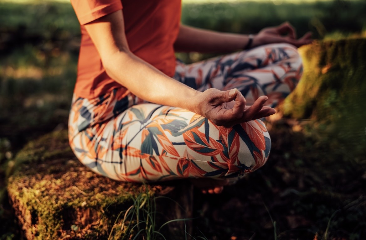 Nahaufnahme: eine Übende sitzt im Schneidersitz auf einem bemoosten Baumstumpf, die Hände in Chin Mudra.
