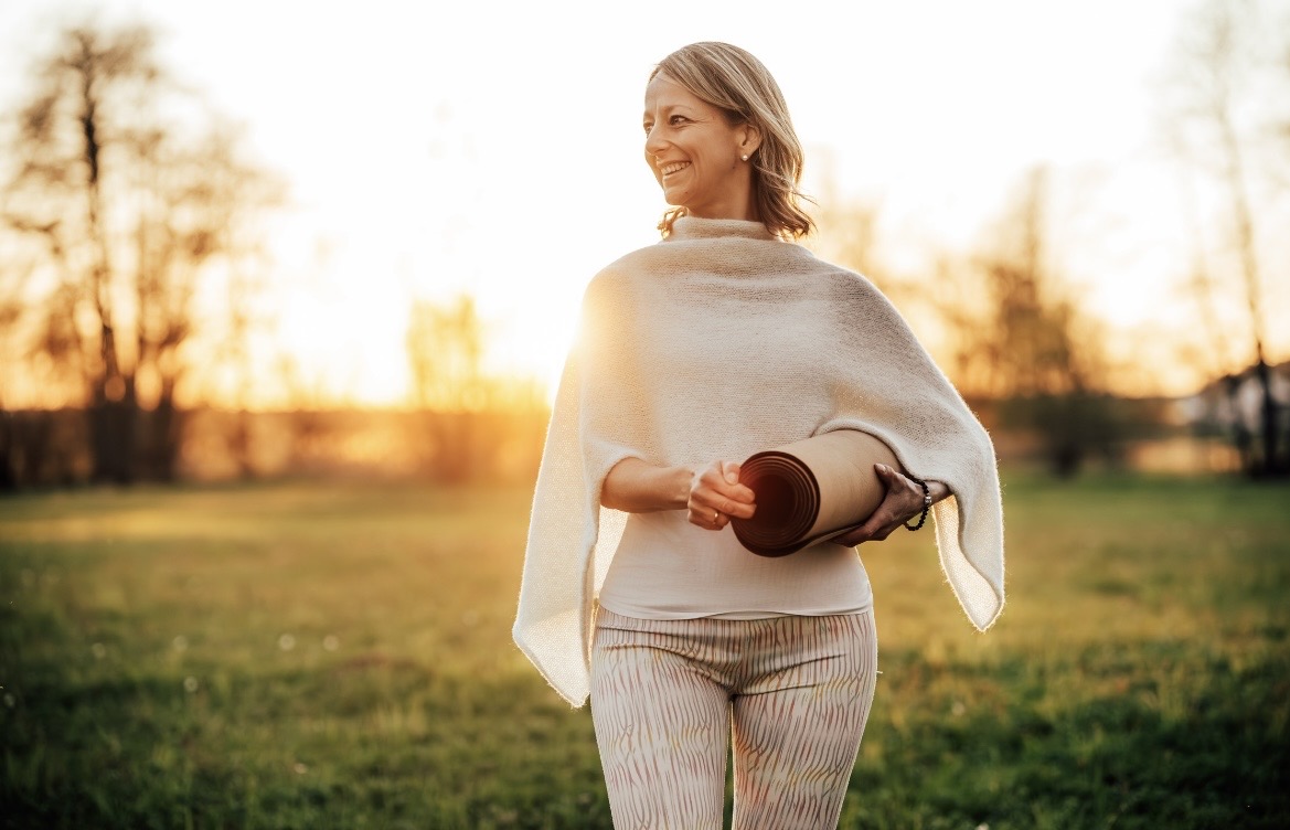 Daniela lacht im Abendlicht, eine Yogamatte unter dem Arm, auf einer Wiese.